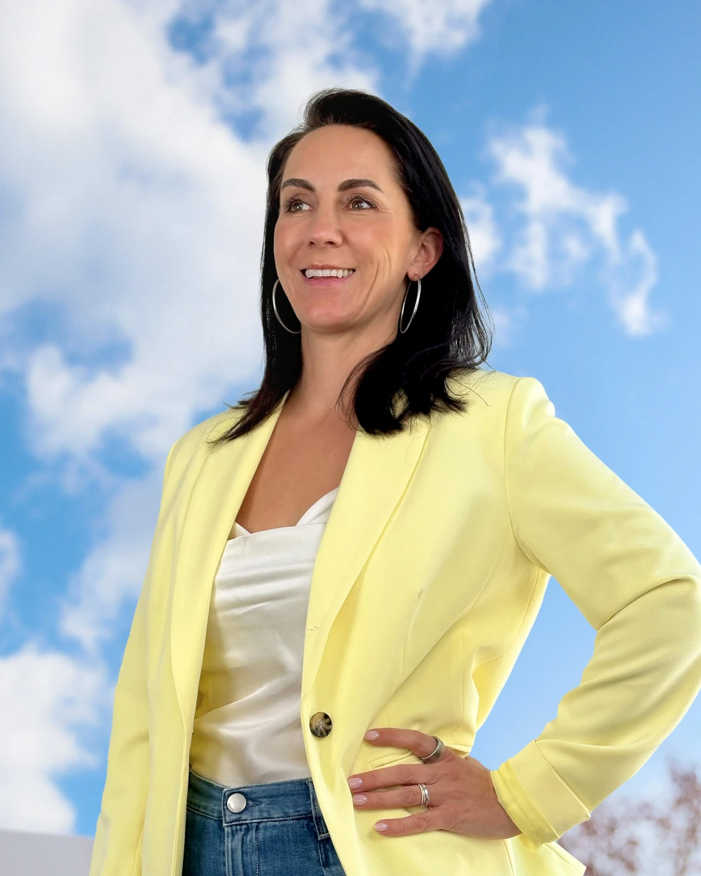 Woman in a yellow blazer standing outdoors in front of a blue sky with clouds looking into the distance