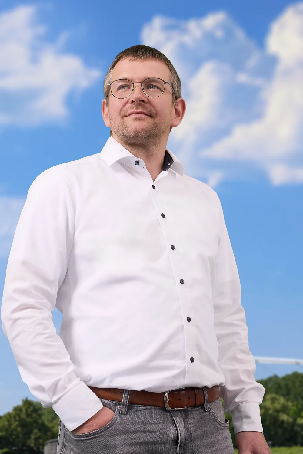 Man in a white shirt and glasses standing outdoors in front of a blue sky with clouds looking into the distance