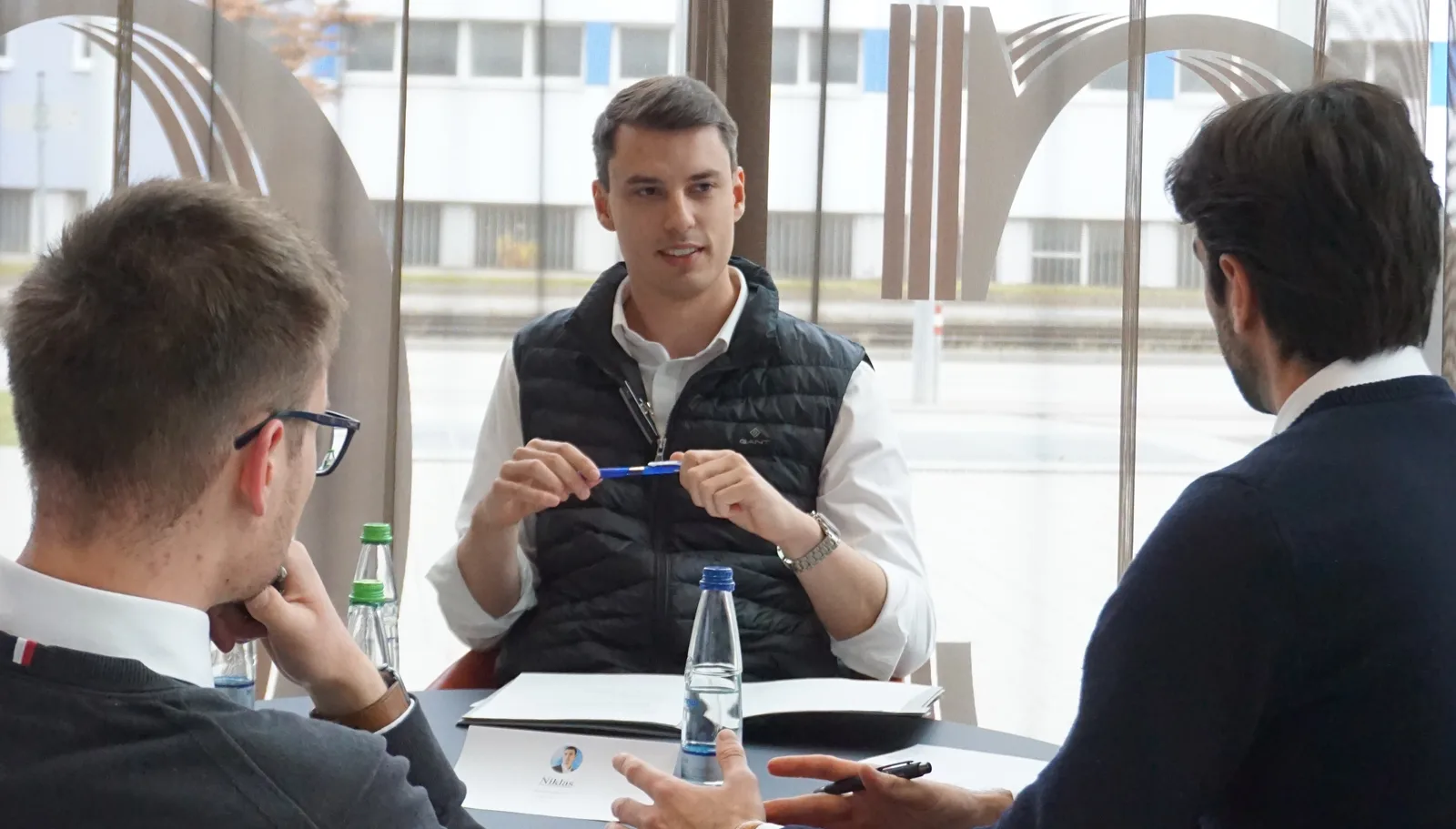 Consultant in a dark vest explaining a case to two participants at a table in front of large windows
