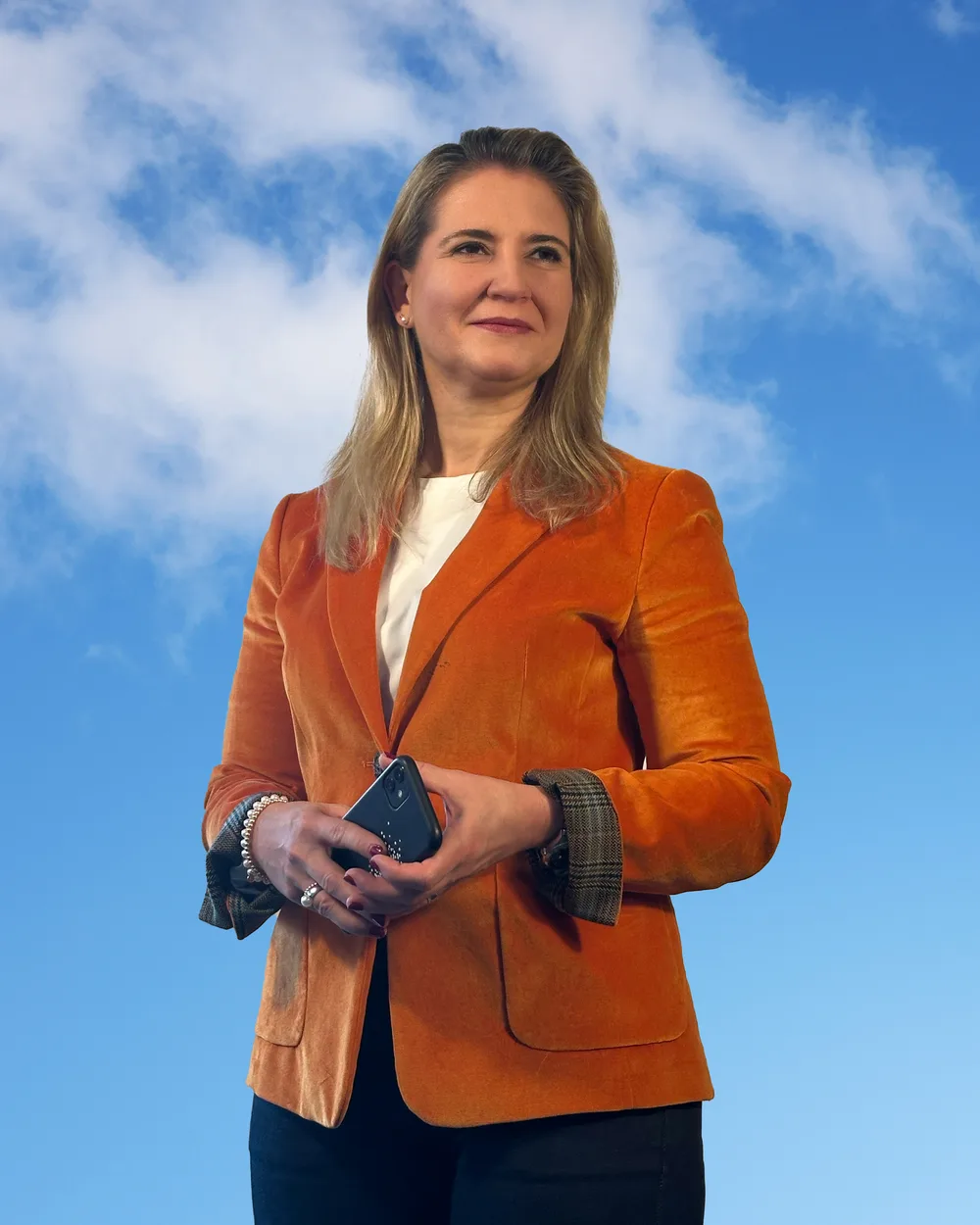 Woman with shoulder-length blonde hair in an orange blazer holding a smartphone in front of a blue sky with clouds
