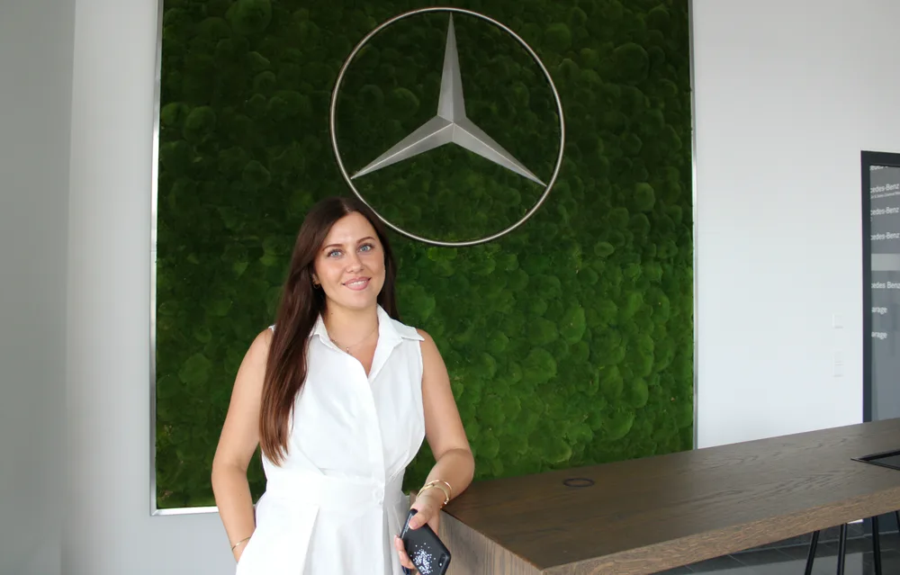 Woman with long brown hair in a white dress standing at a wooden counter in front of a green moss wall with a Mercedes-Benz star